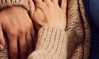 Nahaufnahme der Hände zweier Personen, die in einer unterstützenden Geste zusammen auf einer beigen Strickdecke ruhen - es.