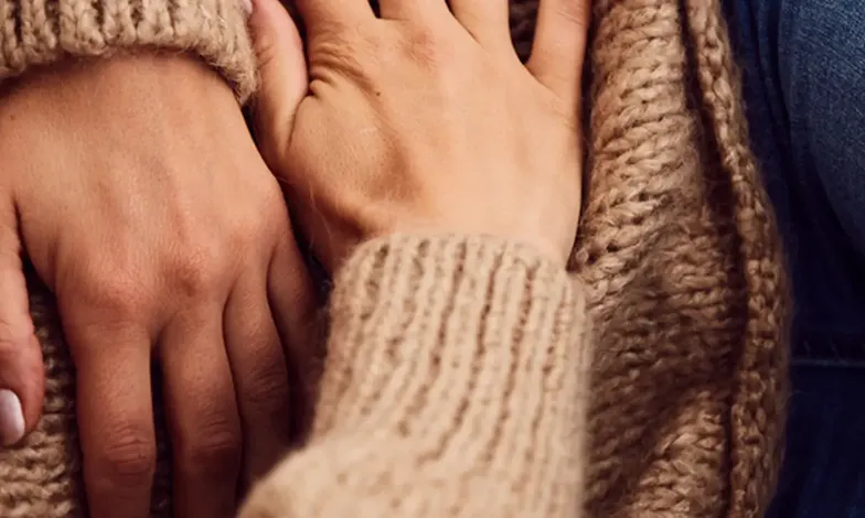 Gros plan sur les mains de deux personnes posées ensemble sur une couverture en tricot beige, dans un geste de soutien - es.